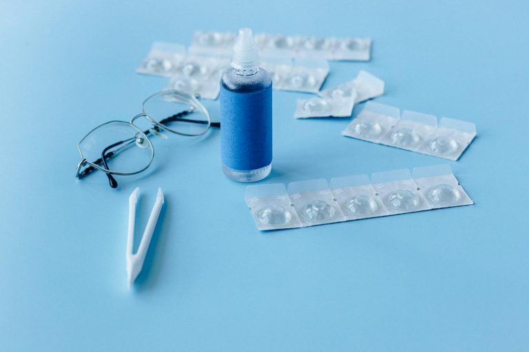 Close-up of contact lens solution bottle with eyeglasses and tweezers on blue backdrop.